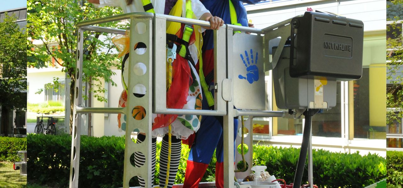 2 Klinikclowns stehen im Korb einer LKW-Arbeitsbühne